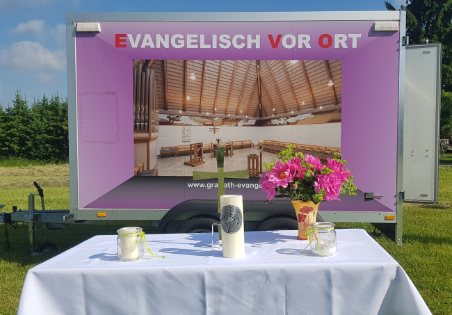 Ein mobiler Hänger mit Kirchenbild steht auf einer Wiese. Davor ist ein Altar aufgebaut.